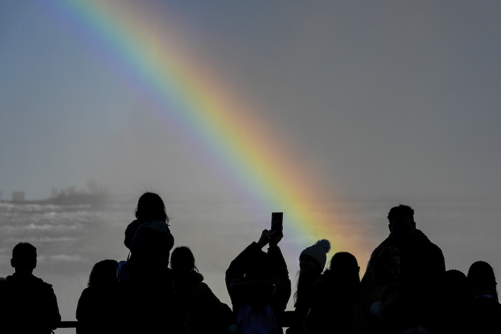 Un gruppo di persone in controluce, fotografate di spalle e a mezzo busto, ammira le cascate e l’arcobaleno