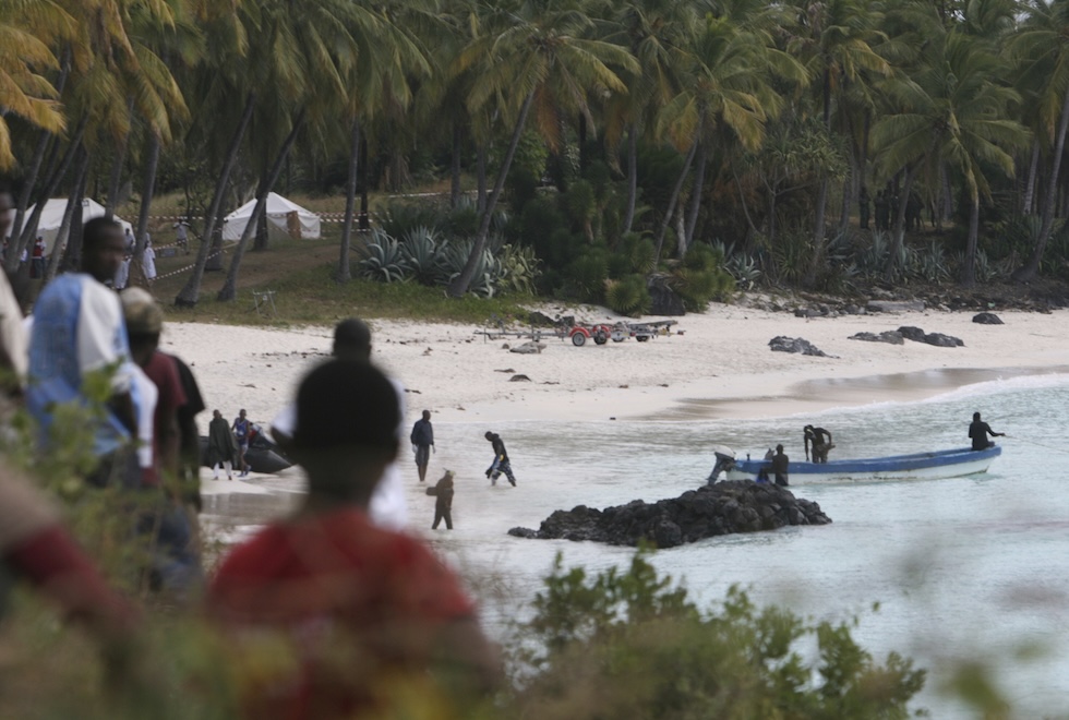 Due persone su una barca e altre su una spiaggia, in una foto scattata da una scogliera vicina