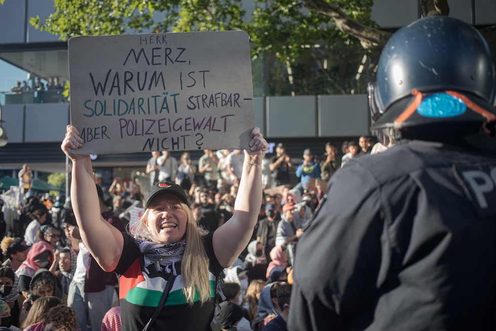 Una manifestazione contro Merz a Berlino, il 2 agosto. Il cartello dice: "Signor Merz, perché viene punita la solidarietà ma non la violenza della polizia?