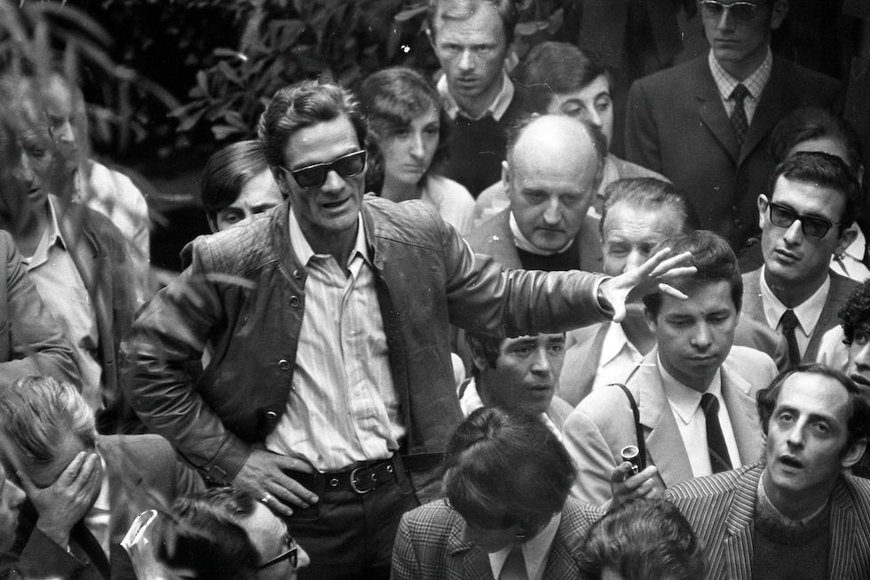 Pasolini in piedi parla e gesticola, da una postazione rialzata, in mezzo a un gruppo di persone