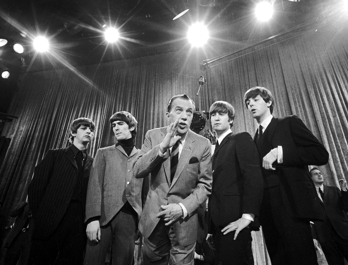 Ed Sullivan, in mezzo ai Beatles, avvicina la mano alla bocca come per dire qualcosa al pubblico