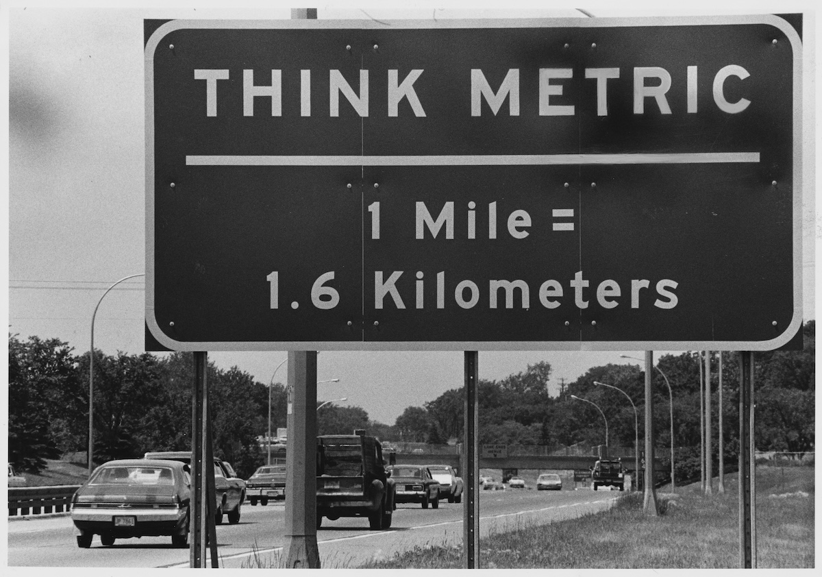 Un cartello stradale vicino al confine con gli Stati Uniti, in Quebec, negli anni Settanta, con la scritta «Pensa in metrico - 1 miglio = 1,6 chilometri»