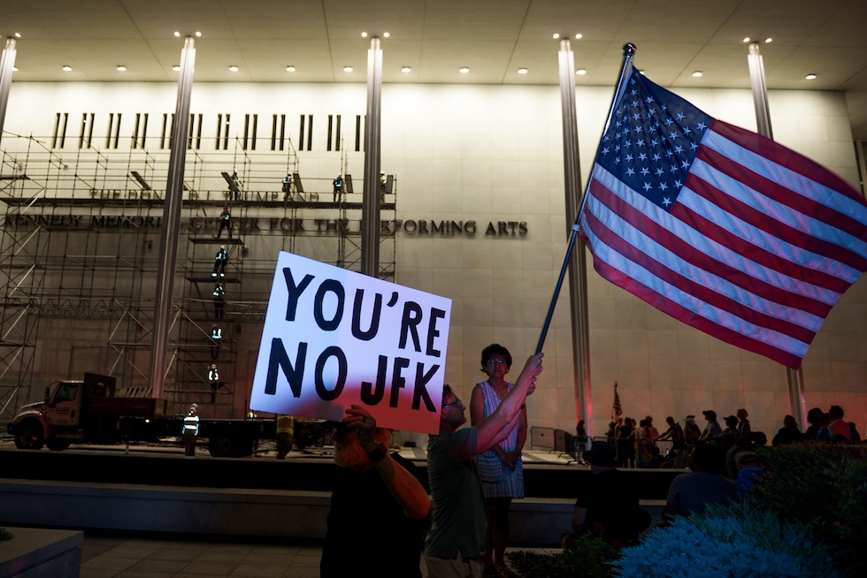 I manifestanti sventolano bandiere statunitensi mentre viene rimossa la scritta con il nome del presidente Donald Trump dal John F. Kennedy Center for the Performing Arts di Washington (AP Photo/Cliff Owen)
