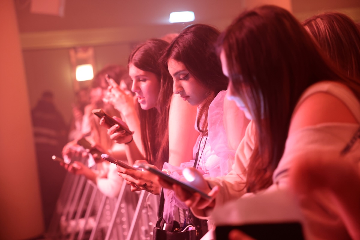 Una fila di ragazze che usano lo smartphone poggiate su una transenna vicino al palco