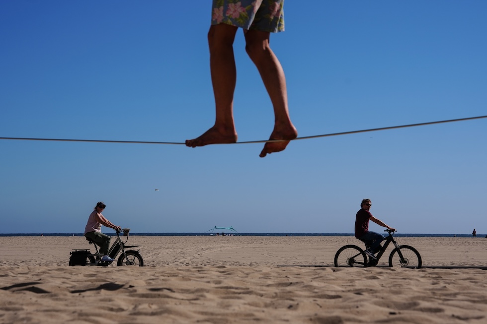 Due persone in bici in spiaggia e un'altra su una slackline, una fettuccia di nylon o poliestere su cui camminare in equilibrio (slackline è anche il nome della disciplina in sè) (AP Photo/Jae C. Hong)