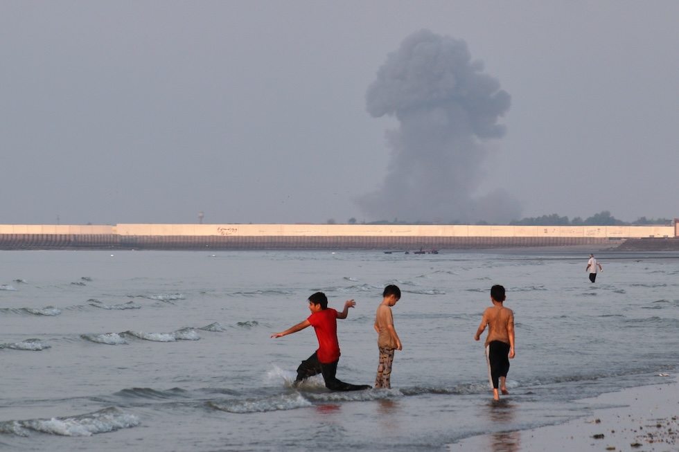 Tre bambini giocano nelle acque dello Stretto di Hormuz, mentre sullo sfondo si vede la colonna di fumo provocata da un'esplosione (Razieh Poudat/ISNA via AP)