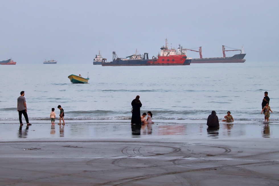 Persone sulla spiaggia, di fronte alle navi commerciali ancorate nello Stretto di Hormuz (Amirhosein Khorgooi/ISNA via AP)