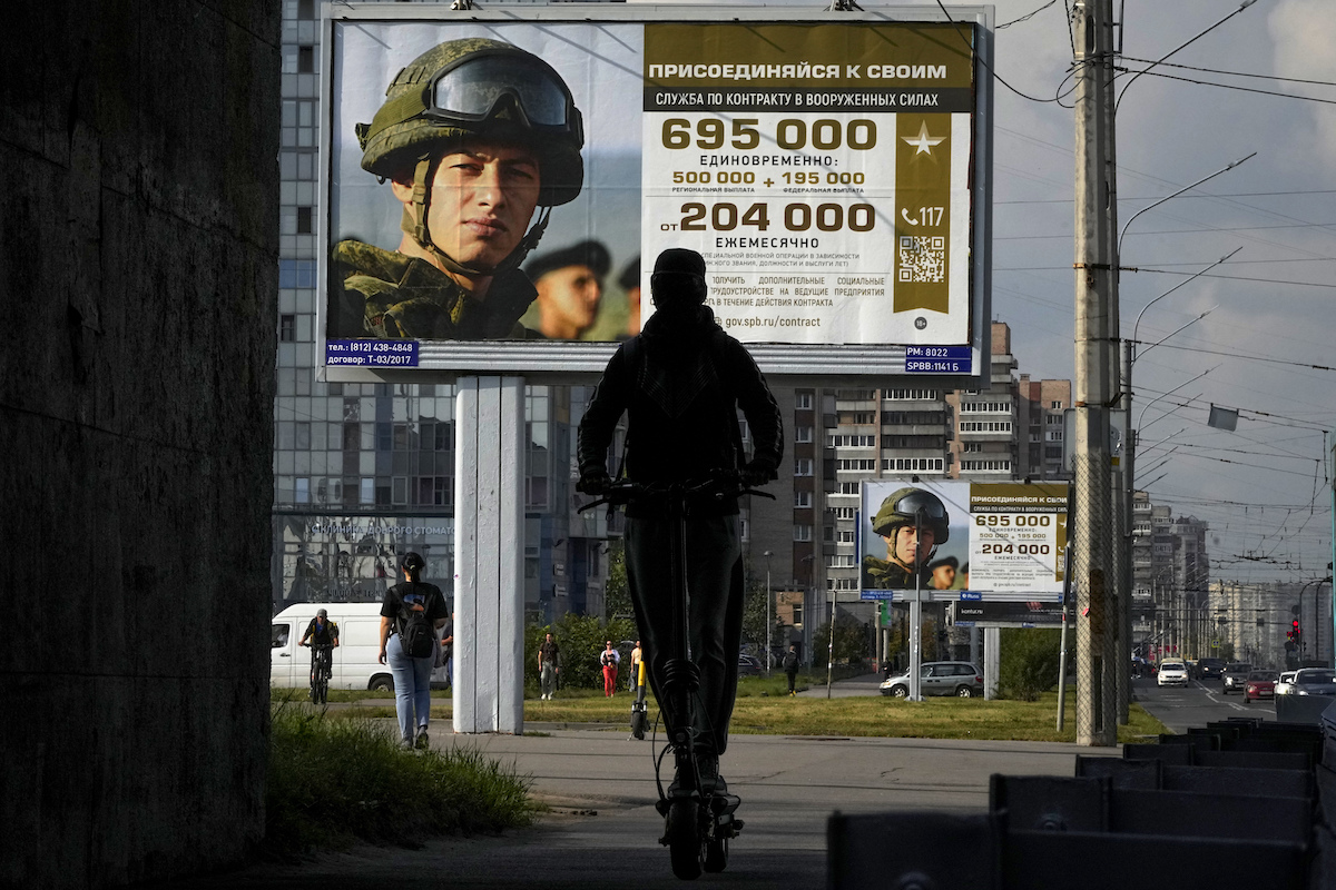 Lavoratori russi in fila per ricevere stipendi in un ufficio, con un manifesto che promuove paghe elevate per i soldati