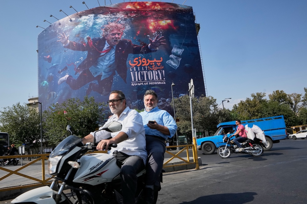 Due motociclisti passano davanti a un cartellone pubblicitario che raffigura il presidente degli Stati Uniti Donald Trump sott’acqua in mezzo a delle esplosioni, con la scritta “Grande vittoria! Stretto di Hormuz” (AP/Vahid Salemi)
