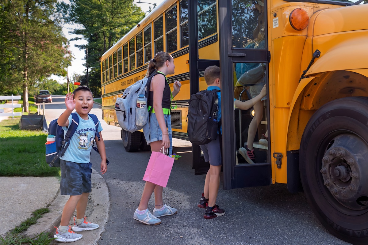 Un bambino saluta con la mano verso l'obiettivo, dietro altri due compagni che stanno salendo sullo scuolabus