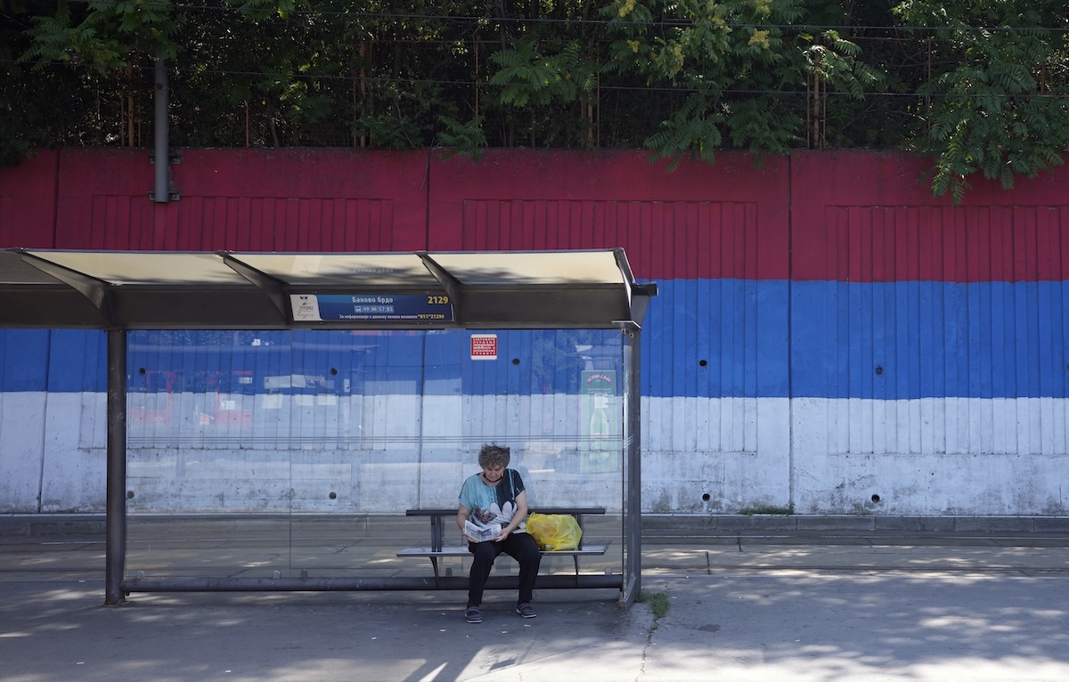 Una donna a una fermata del bus davanti a un muro dipinto con i colori della bandiera serba, a Belgrado (AP/Darko Vojinovic)